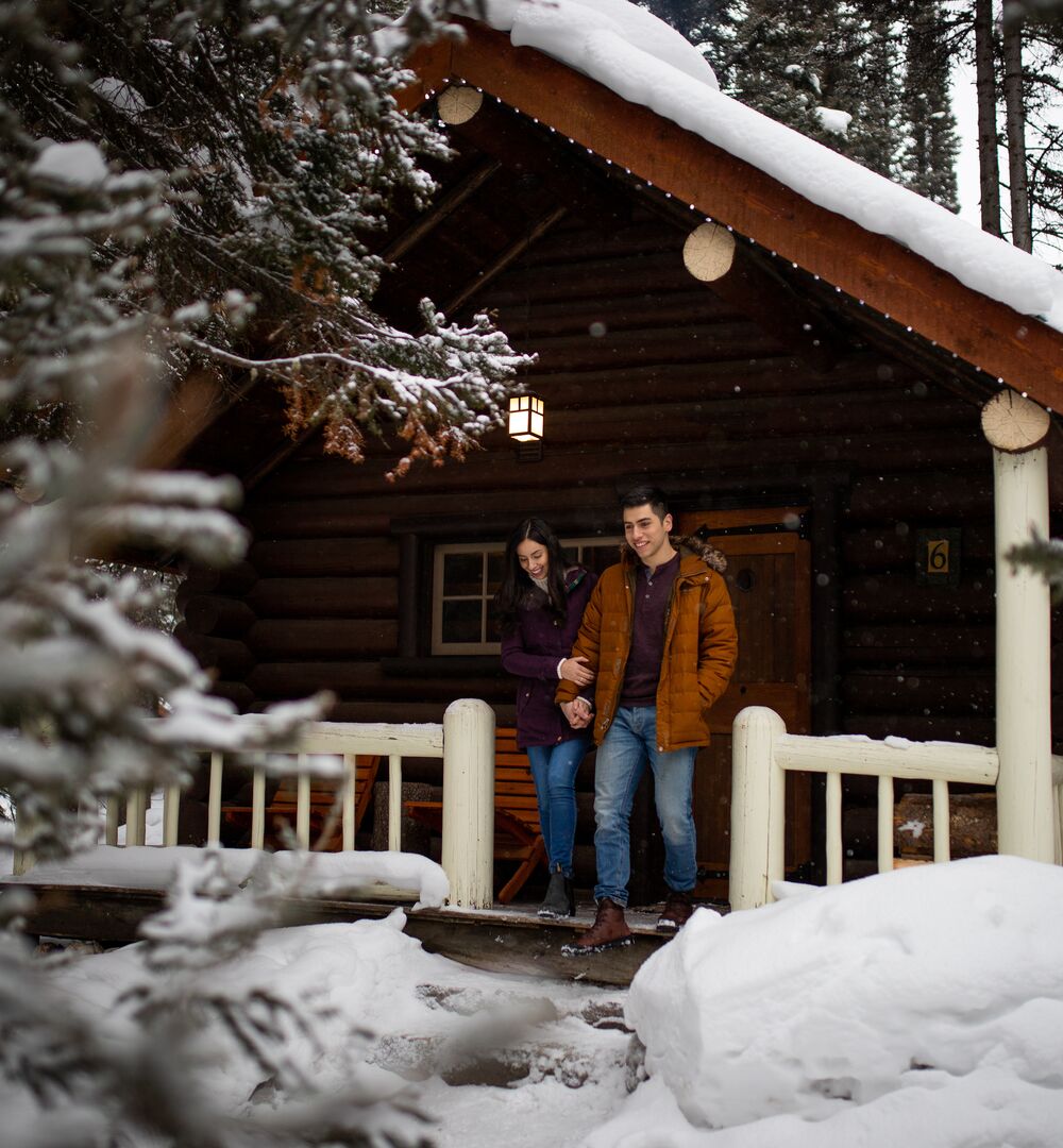 Two people on the front deck of a cabin nestled in a snow covered wooded area